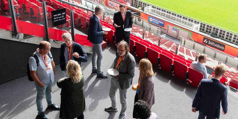 Mensen staan te praten in een kring op de tribune van een voetbalstadion. Op de achtergrond het voetbalveld.